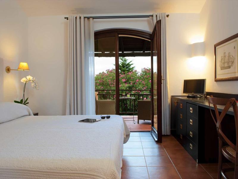 Helles Schlafzimmer mit Doppelbett, Schreibtisch, Stuhl, offenen Balkonfenstern und weißer Wanddekoration.