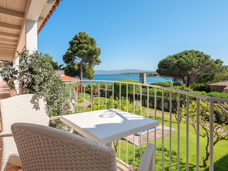Balcon avec table et chaises, vue sur jardin et mer sous un ciel clair.