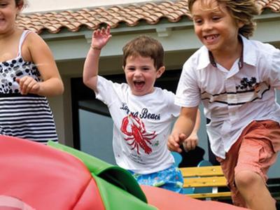 Vier Kinder rennen lachend draußen, vor einem Haus mit roten Dächern, Sommerkleidung, sonniger Tag.