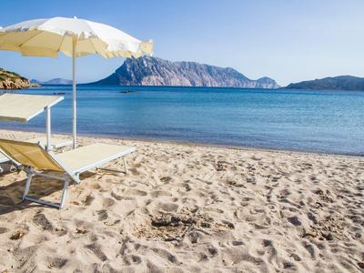 Strand mit Liegestühlen und Sonnenschirm neben klarem, ruhigem Meer und Bergen im Hintergrund.