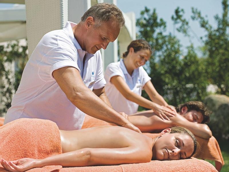 Zwei Personen erhalten Massage von zwei Masseuren im Freien, beide auf orangefarbenen Liegen.