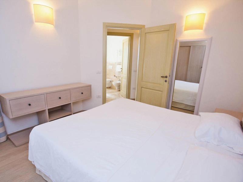 Helles Schlafzimmer mit Doppelbett, Kommode, Spiegel und Blick in ein angrenzendes Bad.