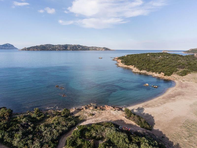 Küste mit klarem Wasser, Sandstrand und bewaldeten Hügeln unter blauem Himmel.