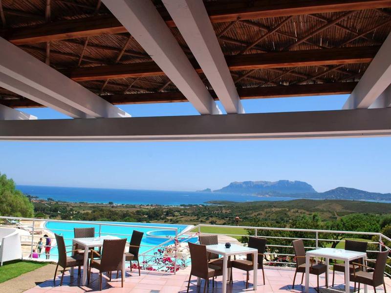 Terrasse d'hôtel avec tables et chaises, vue sur la piscine et la mer avec des montagnes en arrière-plan.