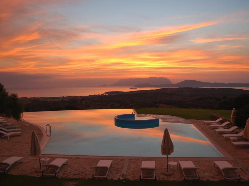 Piscine avec chaises longues et parasols au coucher du soleil avec vue sur montagnes et eau.