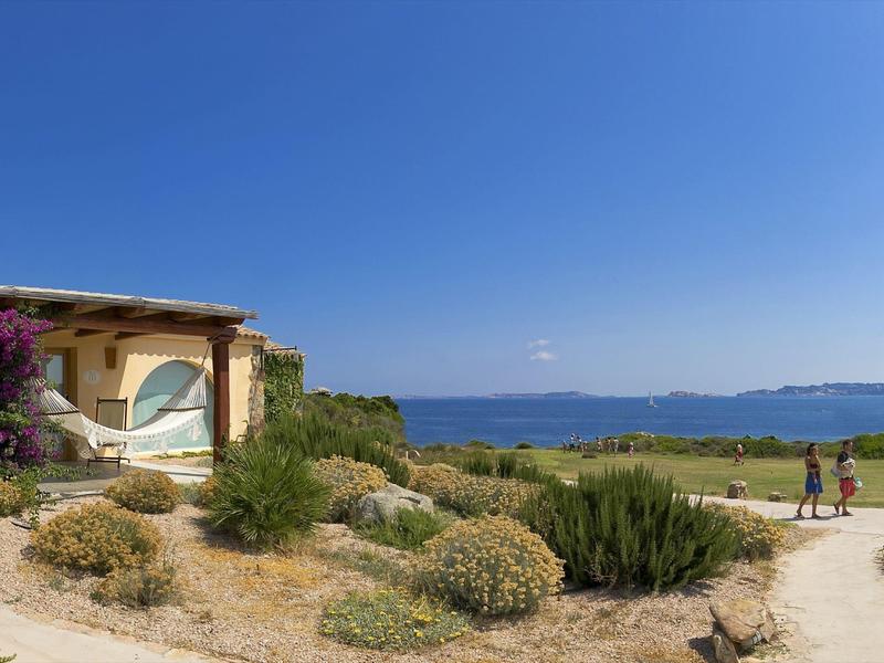 Piccola casa vacanze con amaca accanto a vegetazione mediterranea e vista sul mare.
