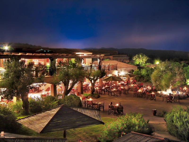 Vista notturna di un hotel illuminato con terrazza esterna e ospiti nel giardino.
