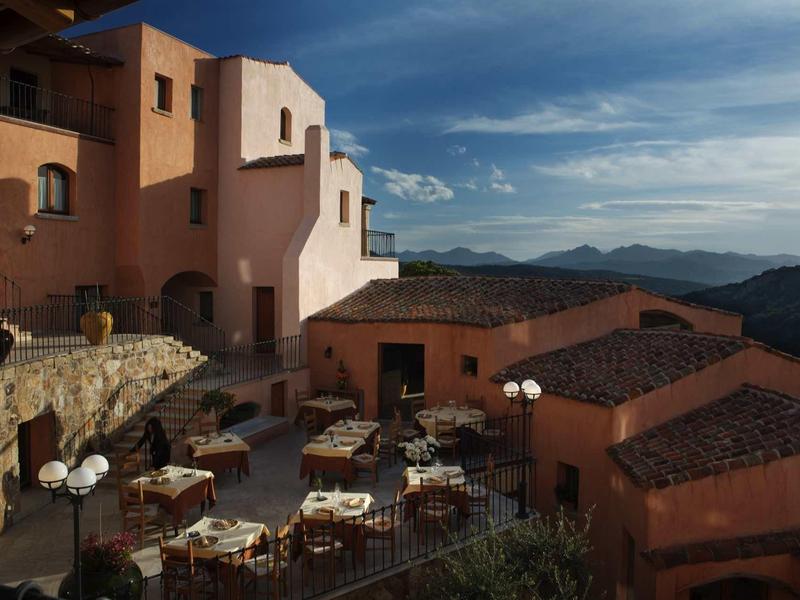 Terras met tafels en stoelen voor een terracottakleurig hotel in bergachtig landschap bij zonsondergang.