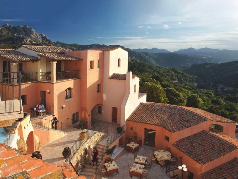 Hotelcomplex met terrassen en tafels ingebed in een bergachtig landschap bij zonnig weer.