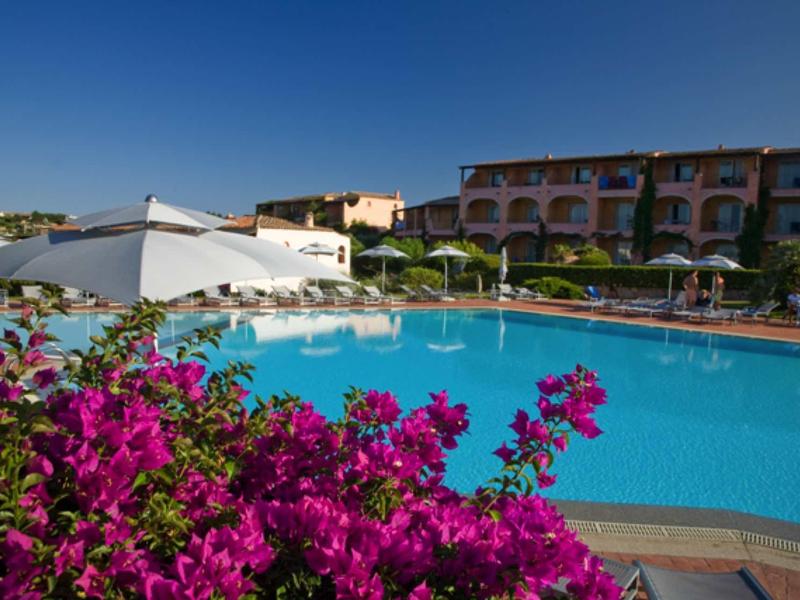 Piscina blu davanti all'hotel con fiori rosa in fiore e ombrelloni in una giornata di sole.
