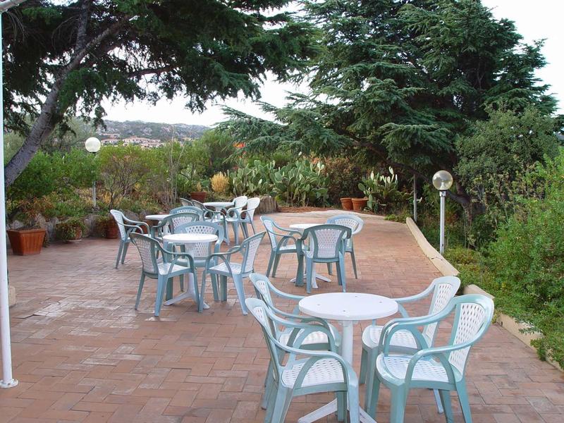 Eine Terrasse mit mehreren weißen Plastiktischen und Stühlen, umgeben von grünen Bäumen und Pflanzen.