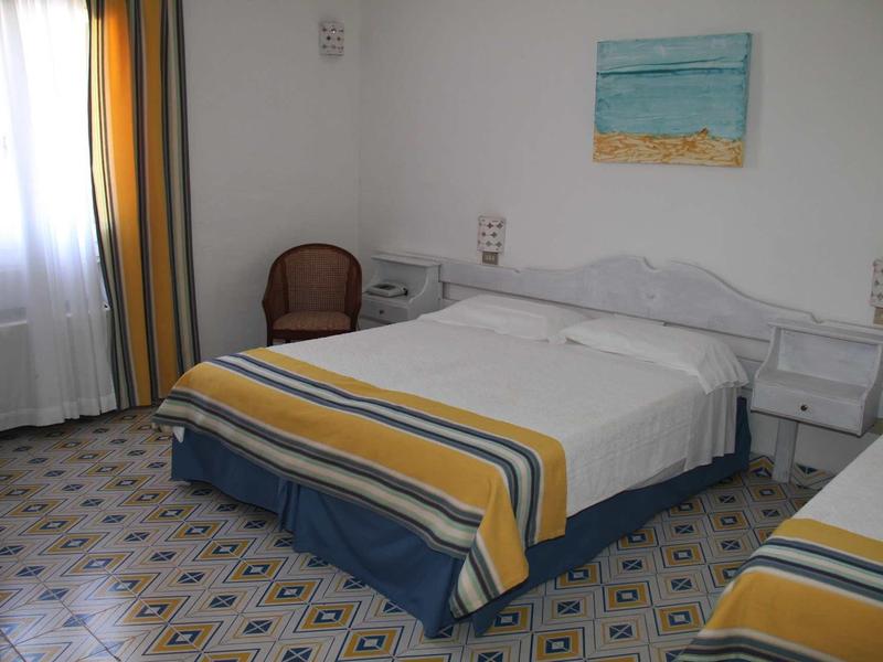 Schlafzimmer mit zwei Betten, gelb-blauen Decken, Fliesenboden und Fenster mit Vorhängen.