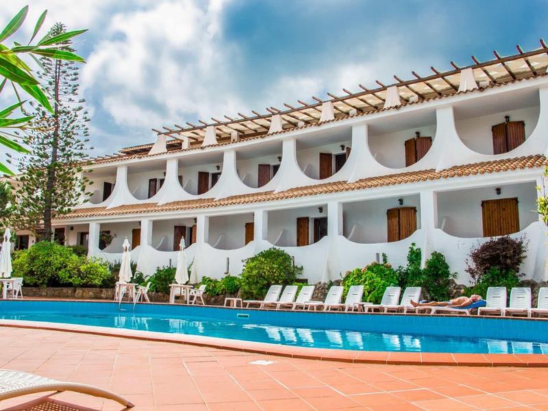 Hotel con edificio bianco, camere con balcone e piscina con lettini in primo piano.