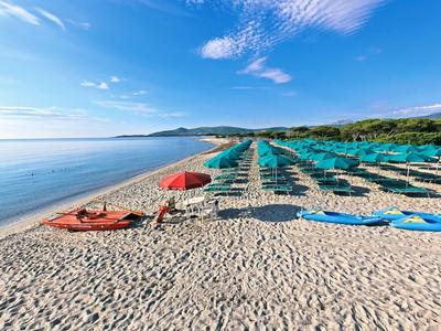 Strand mit feinem Sand, blauen Liegen, grünen Schirmen und ruhigem Wasser unter blauem Himmel.