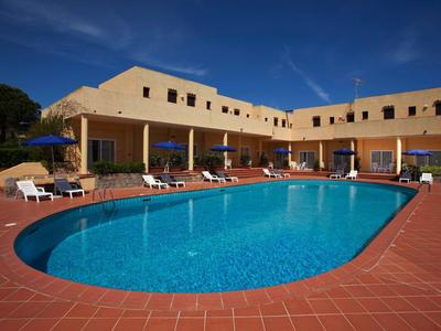 Ein großer, ovaler Pool mit klarem blauem Wasser vor einem gelben Hotelgebäude unter blauem Himmel.