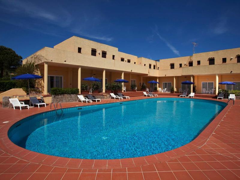 Ein großer, ovaler Pool mit klarem blauem Wasser vor einem gelben Hotelgebäude unter blauem Himmel.