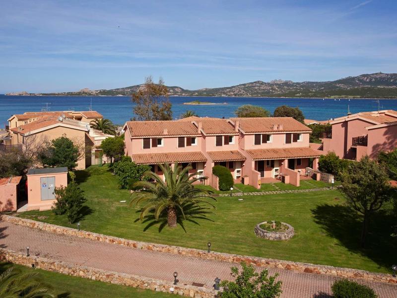 Mehrere rosa Gebäude mit roten Dächern in grüner Landschaft, Meer und Hügel im Hintergrund.