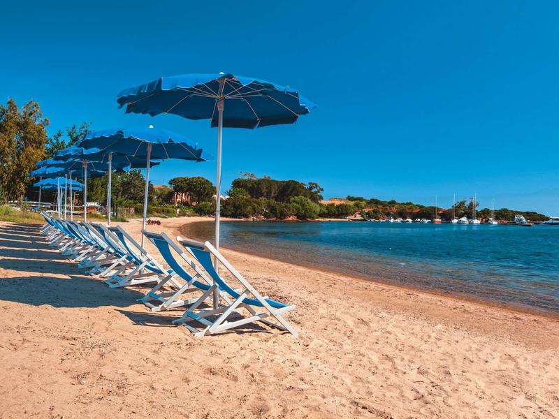 Leere Liegestühle mit blauen Sonnenschirmen stehen am goldenen Sandstrand unter klarem Himmel.