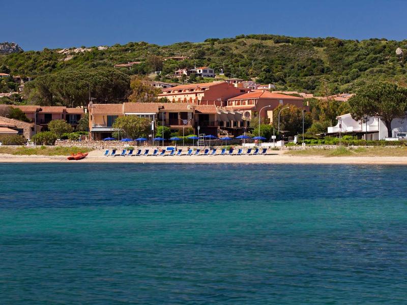 Strandhotel vor grünen Hügeln, Sandstrand mit blauen Liegestühlen, türkisblaues Meer im Vordergrund.