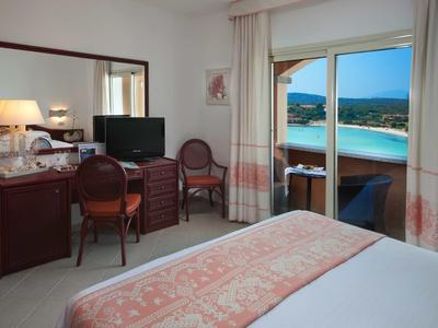 Bright hotel room with double bed, desk, TV, and balcony overlooking the sea.