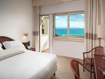 Bright hotel room with balcony and sea view, white bed, and desk with chair.