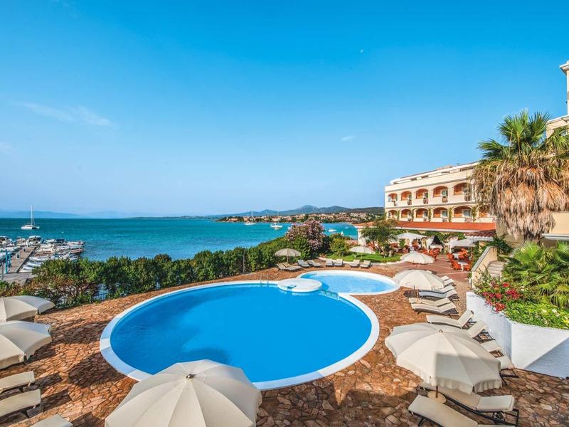 View of hotel pool with umbrellas and sea in the background under a sunny sky.