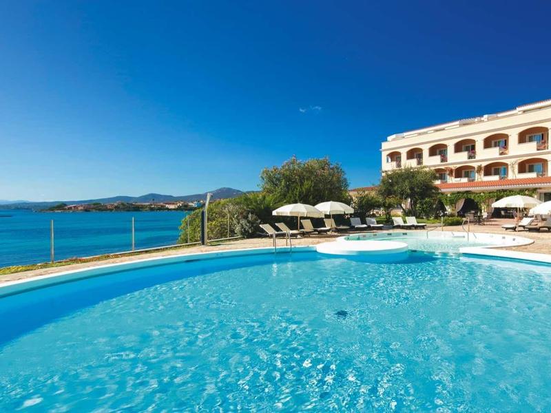 Hotel with large pool, sun umbrellas, and sea view under clear blue sky.