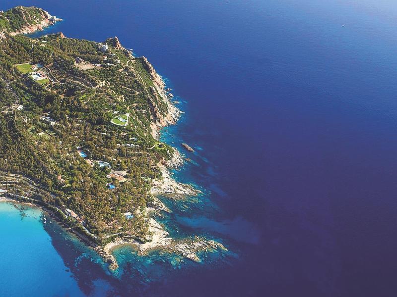 Aerial view of a coastal area with rocky cliffs, clear blue sea, and scattered buildings.