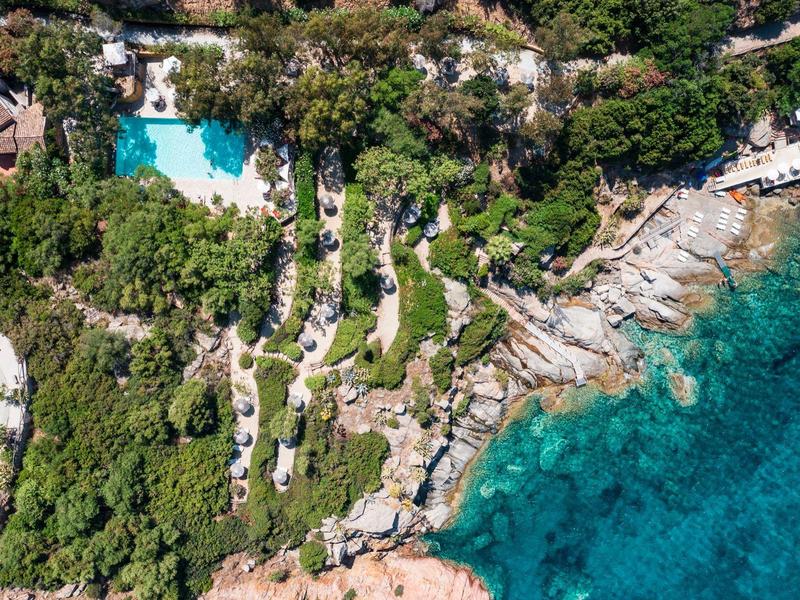 Aerial view of a coastline with clear sea, rocky shore, and a tree-surrounded terrace with a pool.