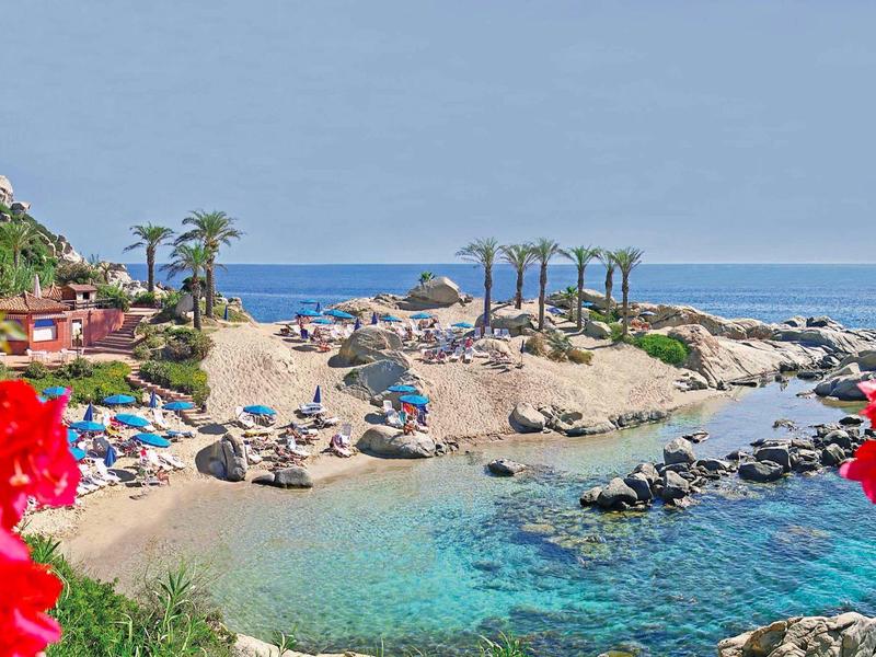 Malá písečná pláž s lehátky mezi skalami a palmami u moře