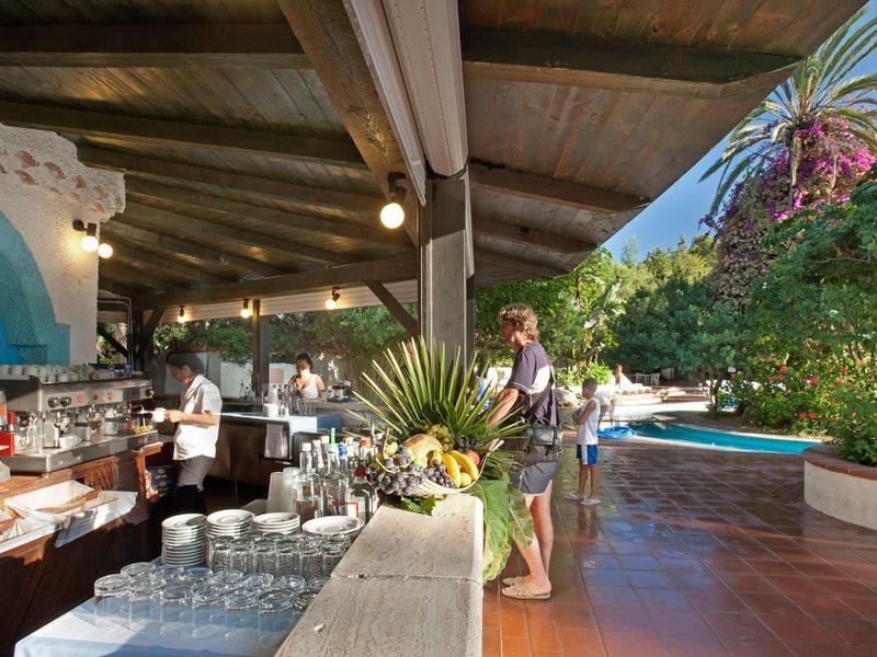 Venkovní bar s dřevěným stropem, hosty, bazénem a tropickou vegetací.