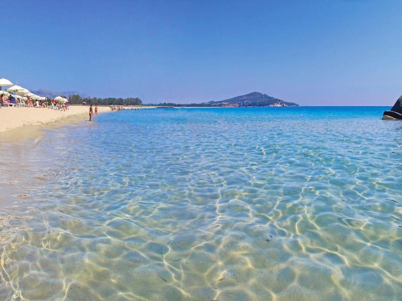 Čistá mělká voda na slunné písčité pláži s deštníky a vzdálenými kopci.