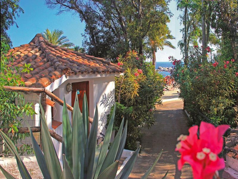 Cesta mezi rostlinami vedoucí k bílému domu s červenou střechou a výhledem na moře