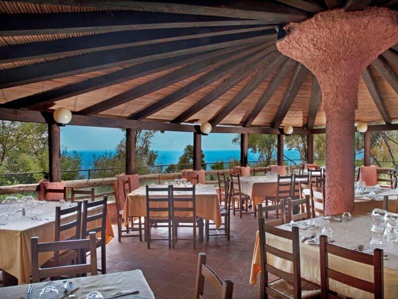 Otevřená restaurace s dřevěnými židlemi, stoly a výhledem na moře pod dřevěnou střechou.