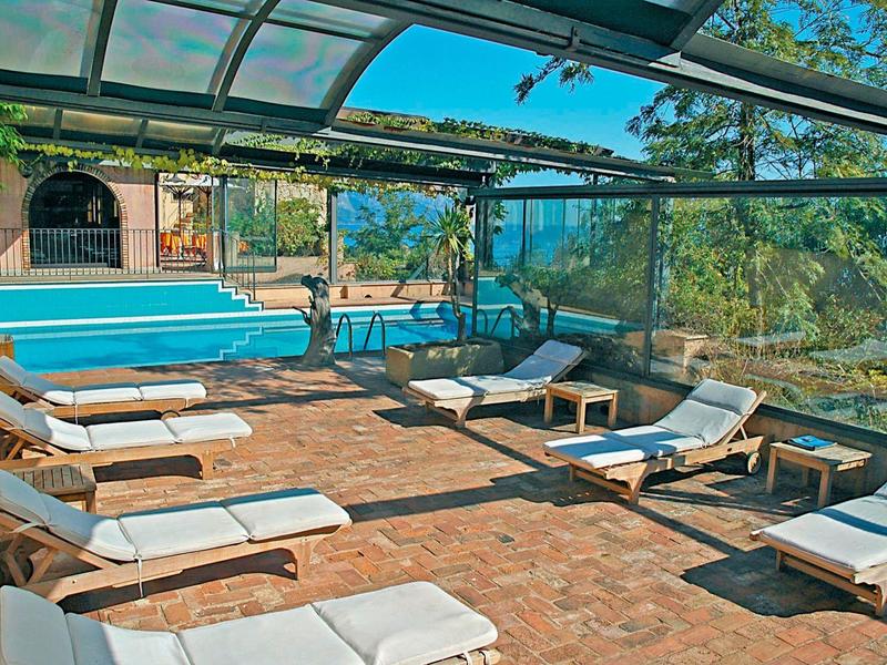 Überdachter Außenpool mit blauer Liegefläche und mehreren weißen Liegestühlen auf rotem Steinboden.
