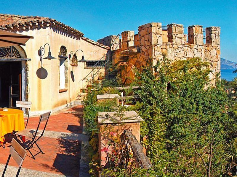 Sonnige Terrasse mit Tischen, Stühlen, mediterranen Gebäuden und weitem Meerblick.
