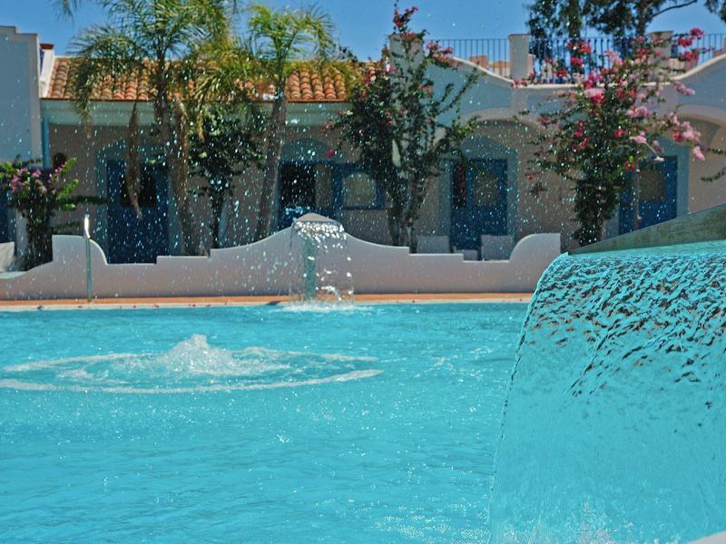 Blaues Wasserbecken mit Springbrunnen, weiße Mauer und Pflanzkübel im Hintergrund.