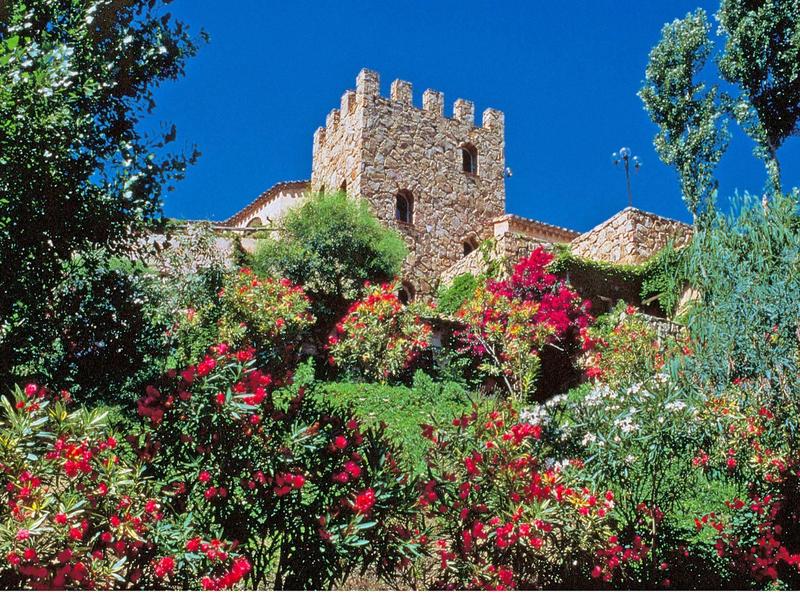 Eine mittelalterliche Steinburg über einem bunt blühenden Garten vor blauem Himmel.
