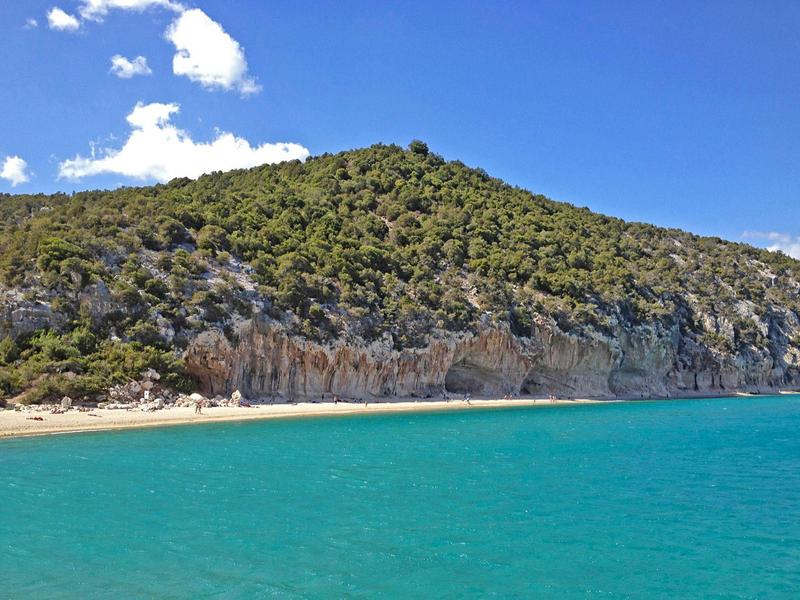Grüner Hügel mit Felsen am Strand, türkisblaues Wasser und blauer Himmel mit Wolken.