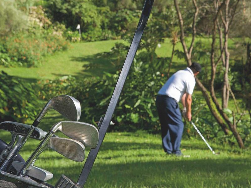 Mann spielt Golf auf grünem Rasen, links stehen zwei leere Klappstühle.