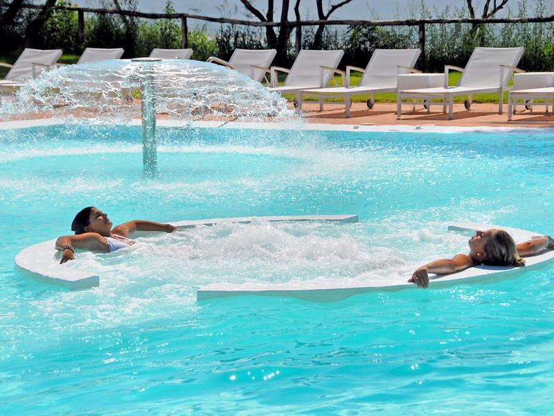 Zwei Personen entspannen auf weißen Wasserliegen in einem klaren blauen Pool mit Wasserfall.