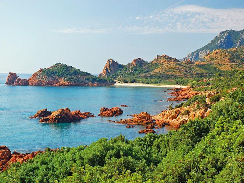 Küstenlandschaft mit rotem Felsen, grünem Gebüsch und blauem Meer unter klarem Himmel
