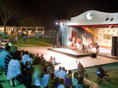 Evening dance performance on a lit outdoor stage with audience seated on chairs and grass.