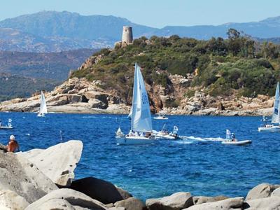 Sailboats sail on blue water near a rocky coast with hills in the background.