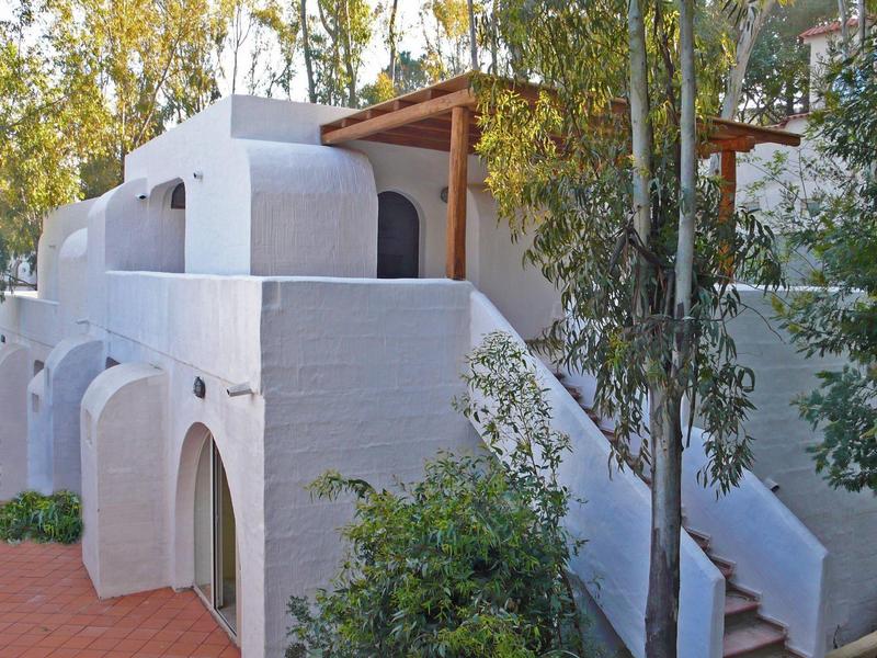 Casa de vacaciones blanca con ventanas arqueadas, porche de madera y escaleras exteriores en ambiente soleado.