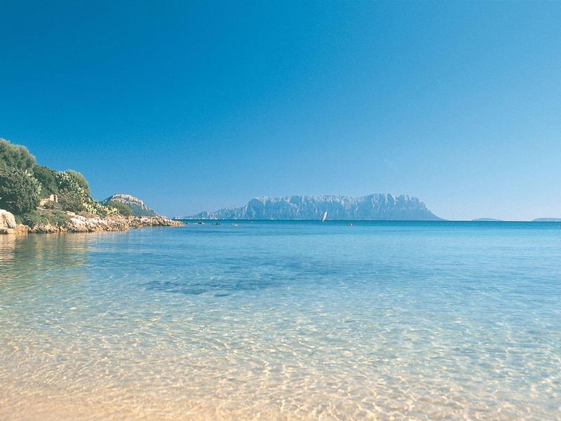 Mare calmo e limpido con costa rocciosa e isola all'orizzonte sotto un cielo azzurro.