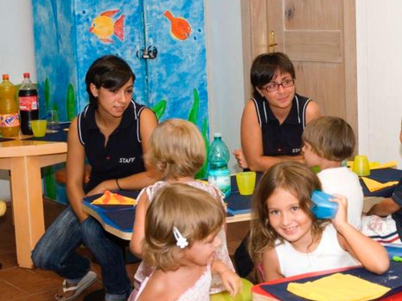 Bambini seduti a un tavolo con assistenti in una stanza luminosa con una tenda colorata.