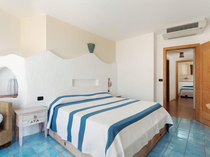 Camera d'hotel luminosa con letto matrimoniale, copriletto a righe blu e bianche, pavimento in piastrelle blu.