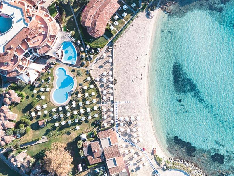Veduta aerea di un resort con piscina, lettini e mare turchese trasparente.