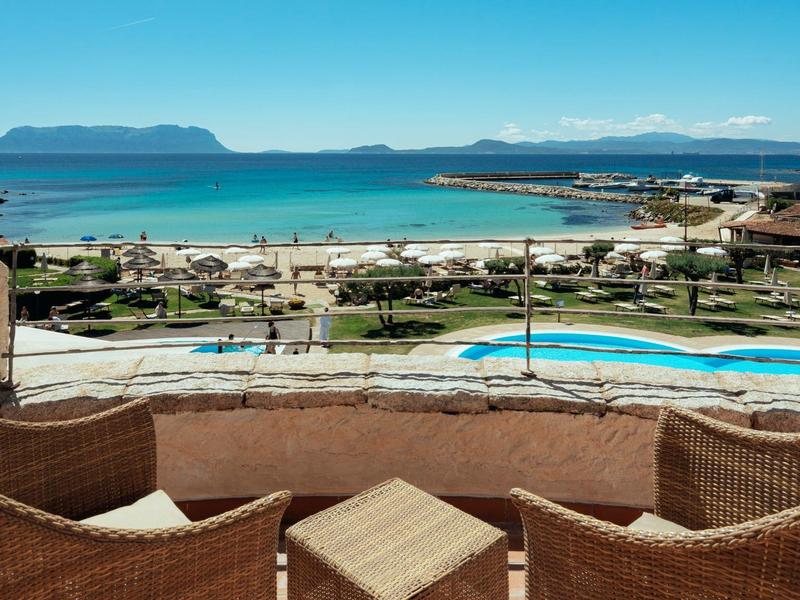 Vista dal balcone con mobili in vimini su piscina, spiaggia e montagne con cielo limpido.